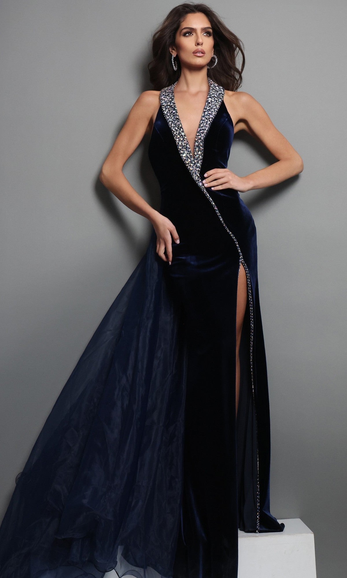 A woman wearing the Long Formal Dress: Jovani 50544 in navy, a sleeveless velvet gown featuring a beaded neckline and high slit, stands against a gray background.